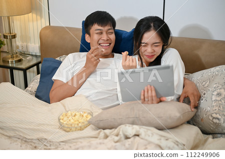 A happy Asian couple are enjoying watching a movie on a tablet and eating popcorn in bed together. 112249906