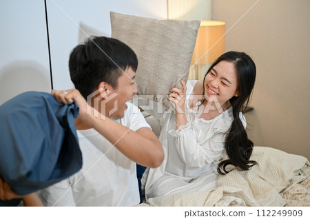A lovely and playful young Asian couple are playing a pillow fight in the bedroom. 112249909