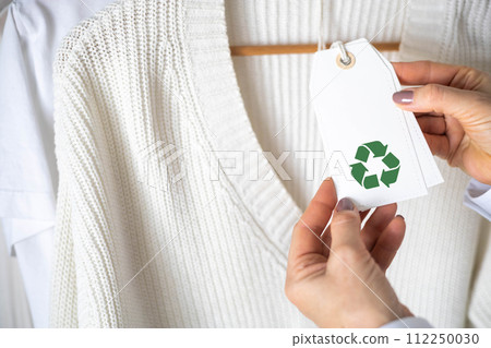 Female hands hold a label from white knitted clothing, which depicts the Recycling sign. 112250030