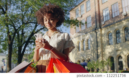 Young biracial woman booking a taxi on smartphone, holding shopping bags 112251736