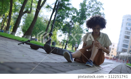 Young woman sitting on ground, rubbing injured knee after electric scooter accident 112251741