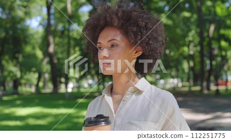 Young thoughtful woman with curly hair drinking coffee outdoors in park 112251775