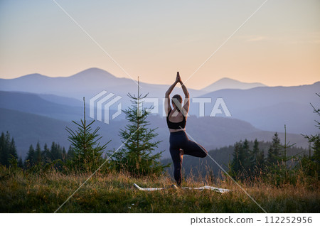 Woman practicing yoga and meditating outdoors in the mountains in a serene, natural setting. Female performing yoga pose on mat, with backdrop of beautiful mountain landscape at sunrise or sunset. Woman practicing yoga and meditating outdoors in the mountains in a serene, natural setting. Female performing yoga pose on mat, with backdrop of beautiful mountain landscape at sunrise or sunset. 112252956