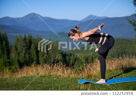 Woman practicing yoga outdoors in the mountains in a serene, natural setting. Female performing yoga pose on blue mat, with backdrop of beautiful mountain landscape at sunrise or sunset. 112252959