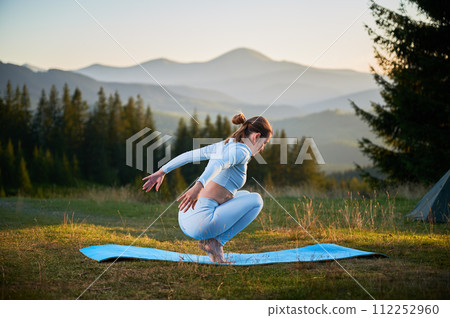 Woman practicing yoga outdoors in the mountains in a serene, natural setting. Female performing yoga pose on blue mat, with backdrop of beautiful mountain landscape at sunrise or sunset. 112252960