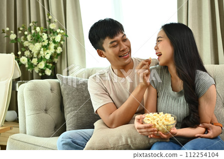 Cheerful young couple having fun while watching TV and eating popcorn at home. Leisure, entertainment and recreation concept 112254104