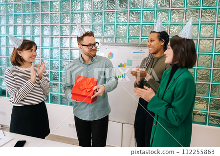 Friendly young multi-ethnic group of employees from startup company celebrating birthday by giving out gift box to happy handsome coworker. Corporate fun celebration in office workplace concept. 112255863