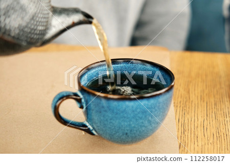 Brewing and Serving Tea. Teapot Pouring Green Tea in Blue Ceramic Cup on Wooden Cafe Table. Two Pottery Handmade Mugs Early in the Morning. Trendy Authentic Porcelain Set. Breakfast Concept. Tea Time 112258017