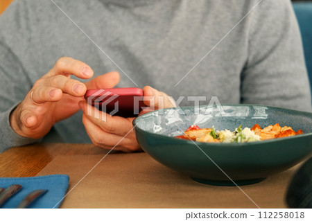 Adult Man in Grey Sweater Set at Wooden Table in Cafe, Using Smartphone. Freelancer Break. Stylish Male Enjoying Lunch and Phone Time in Cozy Restaurant, Eat Caesar Shrimps Salad. Freelancing, Leisure 112258018