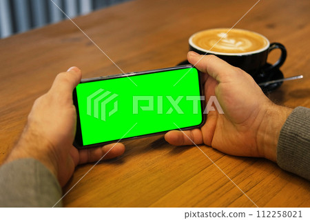 Young man sitting at cafe holding smartphone green mock-up screen in hand. Male person using chroma key mobile phone. Vertical mode. Touching, swiping display, tapping, surfing internet social media 112258021
