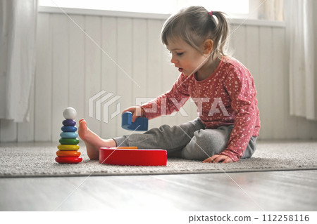 Kid girl play toys at home, kindergarten or nursery. Baby child playing playthings cubes and wood puzzle, pyramid sitting on the floor. Happy preschool education indoor. 3 years old little toddler. 112258116