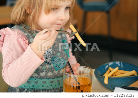 Child Eating with Fork French Fries with Tomato Sauce in Restaurant Indoors. Little Kid Enjoy Unhealthy Fast Food. 3 Years Old Girl Eats Potato Chips at the Table. Little kid having breakfast at cafe. Child Eating with Fork French Fries with Tomato Sauce in Restaurant Indoors. Little Kid Enjoy Unhealthy Fast Food. 3 Years Old Girl Eats Potato Chips at the Table. Little kid having breakfast at cafe. 112258122