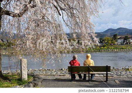 日本春天的京都,嵐山週邊的街道,旅遊勝地桂川,櫻花盛開。一對老夫婦坐在長椅上賞櫻花。 日本春天的京都,嵐山週邊的街道,旅遊勝地桂川,櫻花盛開。一對老夫婦坐在長椅上賞櫻花。 112258794