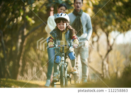 little asian girl riding bike in city park with parents in background little asian girl riding bike in city park with parents in background 112258863