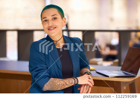 Tattooed woman with electric blue hair sits at desk with laptop Tattooed woman with electric blue hair sits at desk with laptop 112259169