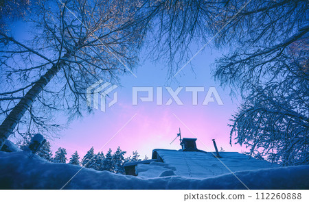 Winter landscape in the evening. View through the trees to a snowy roof with a chimney. Village in the forest in snowy winter at sunset Winter landscape in the evening. View through the trees to a snowy roof with a chimney. Village in the forest in snowy winter at sunset 112260888
