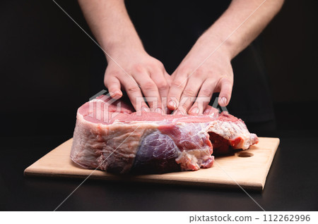 man massages a fresh piece of beef with his hands on a wooden cutting board. Close up on black background 112262996