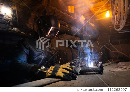 Young mechanic with a welding machine in an old dirty garage at night. Man in protection doing welding work, hobby 112262997