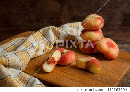 Cutting board with halves and several whole saturn peaches or flat peaches on wooden background with yellow kitchen towel.. Cutting board with halves and several whole saturn peaches or flat peaches on wooden background with yellow kitchen towel.. 112263294