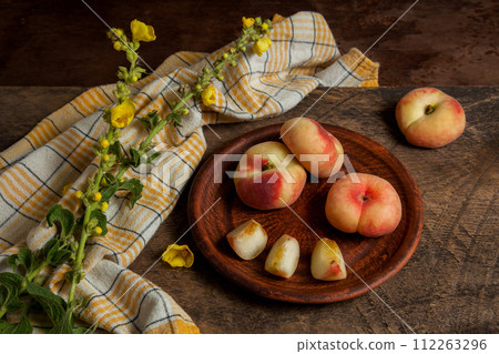 Clay plate with cut and whole saturn peaches or flat peaches on wooden background with yellow kitchen towel.. Clay plate with cut and whole saturn peaches or flat peaches on wooden background with yellow kitchen towel.. 112263296
