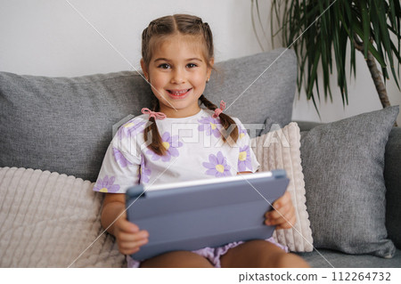 Cute little girl hold tablet in hands and look into camera. Girl sitting on sofa at home 112264732