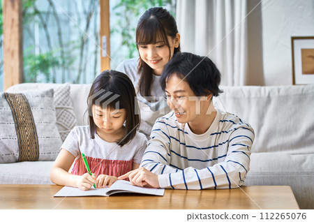 A young father teaching his daughter and her mother watching over her in the living room A young father teaching his daughter and her mother watching over her in the living room 112265076