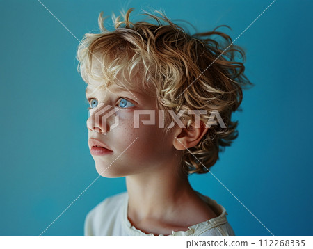 Young boy with blonde curly hair and blue eyes stands by blue wall 112268335