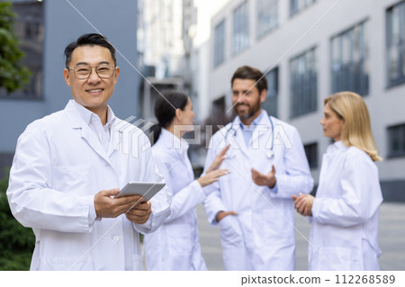 Confident Asian male doctor holding a tablet smiles at camera with medical team engaged in discussion behind him. Confident Asian male doctor holding a tablet smiles at camera with medical team engaged in discussion behind him. 112268589