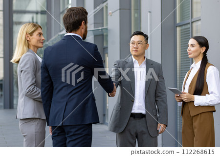 Ethnically diverse business professionals shaking hands and greeting each other outside a modern office building. 112268615