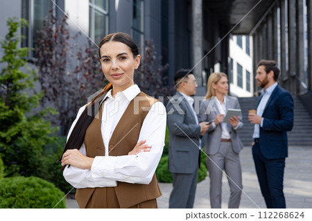 Professional woman in smart casual attire confidently posing with crossed arms. Colleagues conversing in the backdrop. 112268624