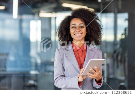 Portrait of a smiling and successful young African American woman in a suit standing in a modern office, holding a tablet and looking at the camera. Portrait of a smiling and successful young African American woman in a suit standing in a modern office, holding a tablet and looking at the camera. 112268631