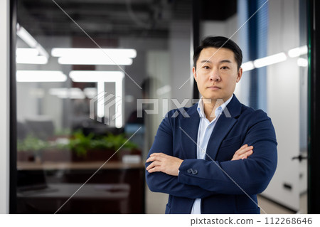 Portrait of a confident and successful young Asian man standing in the office in a suit and looking seriously at the camera with his arms crossed on his chest. Portrait of a confident and successful young Asian man standing in the office in a suit and looking seriously at the camera with his arms crossed on his chest. 112268646