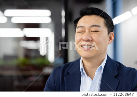 Close-up photo of Asian young man in suit sitting in office with closed eyes and smiling resting, relaxing. Close-up photo of Asian young man in suit sitting in office with closed eyes and smiling resting, relaxing. 112268647