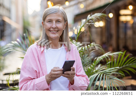 Portrait of a smiling senior woman in a pink shirt standing outside in the middle of the city and using a mobile phone, looking confidently at the camera. Portrait of a smiling senior woman in a pink shirt standing outside in the middle of the city and using a mobile phone, looking confidently at the camera. 112268651