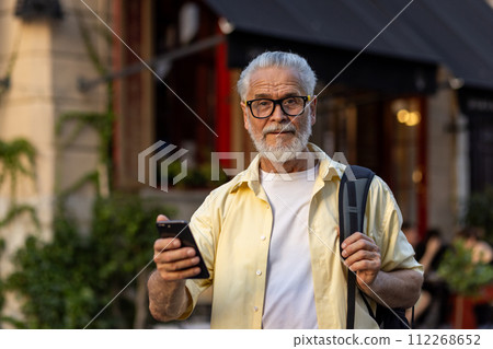 Portrait of a gray-haired senior man dressed in casual clothes and glasses, holding a backpack and a mobile phone, standing on a city street and looking at the camera with a smile. 112268652