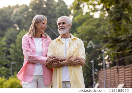 Senior happy couple, gray-haired woman and man in casual clothes holding hands and hugging, walking in the city, smiling, talking to each other. Senior happy couple, gray-haired woman and man in casual clothes holding hands and hugging, walking in the city, smiling, talking to each other. 112268653