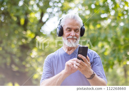 Close-up photo of senior smiling gray-haired man standing, walking in park, doing sports in headphones and chatting on mobile phone. Close-up photo of senior smiling gray-haired man standing, walking in park, doing sports in headphones and chatting on mobile phone. 112268660