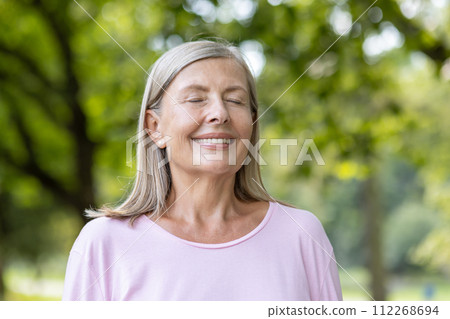 Close-up portrait of smiling senior gray-haired woman in pink t-shirt standing in park and resting, breathing relaxed with eyes closed after walking and doing sports. 112268694