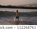 Young boy in blue shorts running and splashing in ocean waves at dusk, with rocks in background. Boy Splashing Through Ocean Waves. 112268761