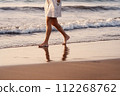 An elderly woman in a dress walks alone on the beach at sunset, reflecting on the wet sand. Senior Woman Walking Barefoot on Beach. 112268762