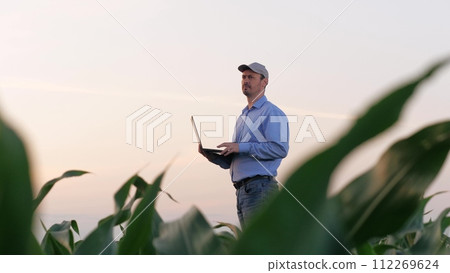 Skilled modern farmer with laptop examines corn growing in rural field. Careful agronomist looks at corn plant holding laptop. Countryman expert works on laptop inspecting corn field in summer Skilled modern farmer with laptop examines corn growing in rural field. Careful agronomist looks at corn plant holding laptop. Countryman expert works on laptop inspecting corn field in summer 112269624