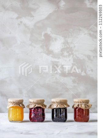 AI-generated content. Four colorful glass jars with fabric lids and jam of different flavor and color on marble surface and white background. High quality photo 112269803