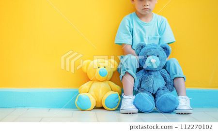AI-generated content. A sad boy in blue T-shirt sits against an empty yellow wall, next to his favorite multi-colored yellow and blue teddy bears. Children health concept, World Autism Awareness Day 112270102