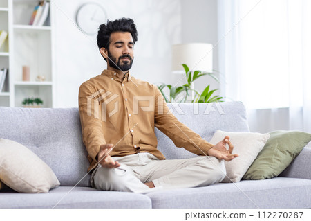 Calm middle eastern man sitting on pillowed sofa in siddhasana position and using hands mudra with closed eyes. Calm guy breathing deeply and practising yoga at home for reducing stress levels. Calm middle eastern man sitting on pillowed sofa in siddhasana position and using hands mudra with closed eyes. Calm guy breathing deeply and practising yoga at home for reducing stress levels. 112270287