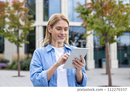 Smiling blonde woman using digital tablet while standing outdoors at pedestrian space with trees. Confident landscape designer developing green zone area in district with futuristic skyscrapers. 112270337