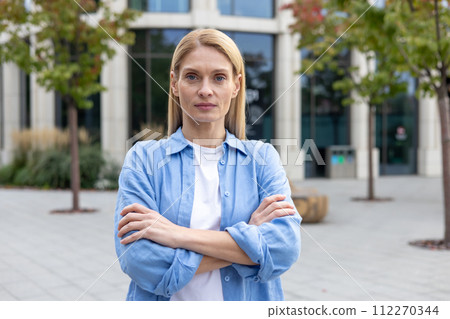 Serious blonde woman in blue shirt standing alone near glass corporate building with hands folded on chest. Successful business owner feeling confident in career development and opportunities. 112270344
