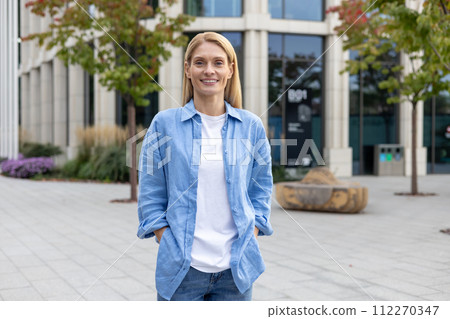 Relaxed millennial woman in stylish casual outfit holding hands in pockets while looking at camera with sincere smile. Optimistic lady spending free day while walking calmly along alley with shops. 112270347