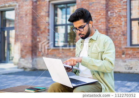 Young indian man in olive shirt sitting on bench at campus with pc on laps and wired earphones. Concentrated student pointing with finger at laptop screen while talking with group mate in online call. Young indian man in olive shirt sitting on bench at campus with pc on laps and wired earphones. Concentrated student pointing with finger at laptop screen while talking with group mate in online call. 112270352