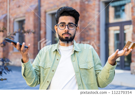 Portrait of uncertain man in eyewear spreading hands in confusion while holding modern smartphone outside. Unhappy male feeling displeased with loosing connection on gadget with technical problems. Portrait of uncertain man in eyewear spreading hands in confusion while holding modern smartphone outside. Unhappy male feeling displeased with loosing connection on gadget with technical problems. 112270354