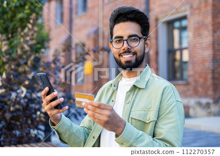 Young arab man holding mobile phone and credit card while looking at camera with wide smile. Cheery bearded guy confirming online cashless payment in banking application during walk outside. Young arab man holding mobile phone and credit card while looking at camera with wide smile. Cheery bearded guy confirming online cashless payment in banking application during walk outside. 112270357